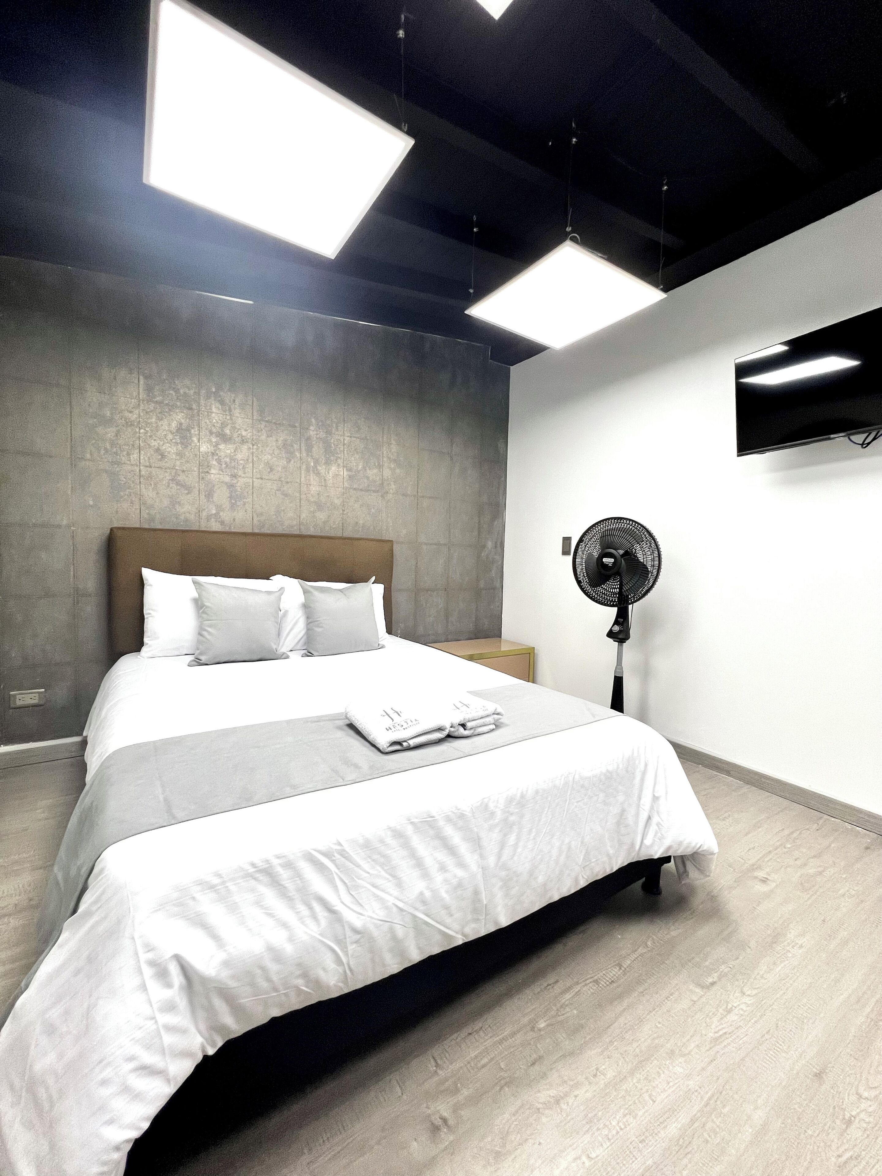 A modern hotel room with a large bed, a flat-screen TV mounted on the wall, and a standing fan.