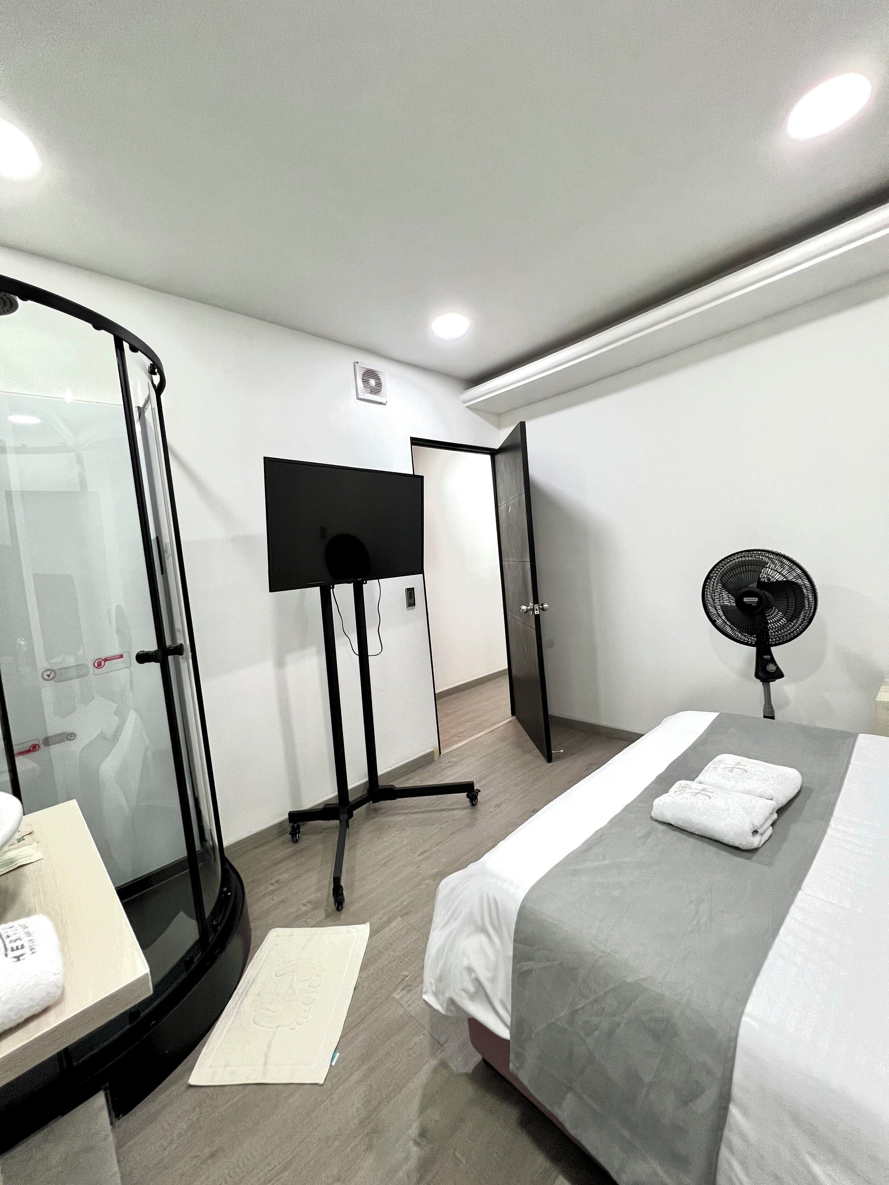A hotel room with a bed, a desk, a glass shower enclosure, a standing fan, and a wardrobe.