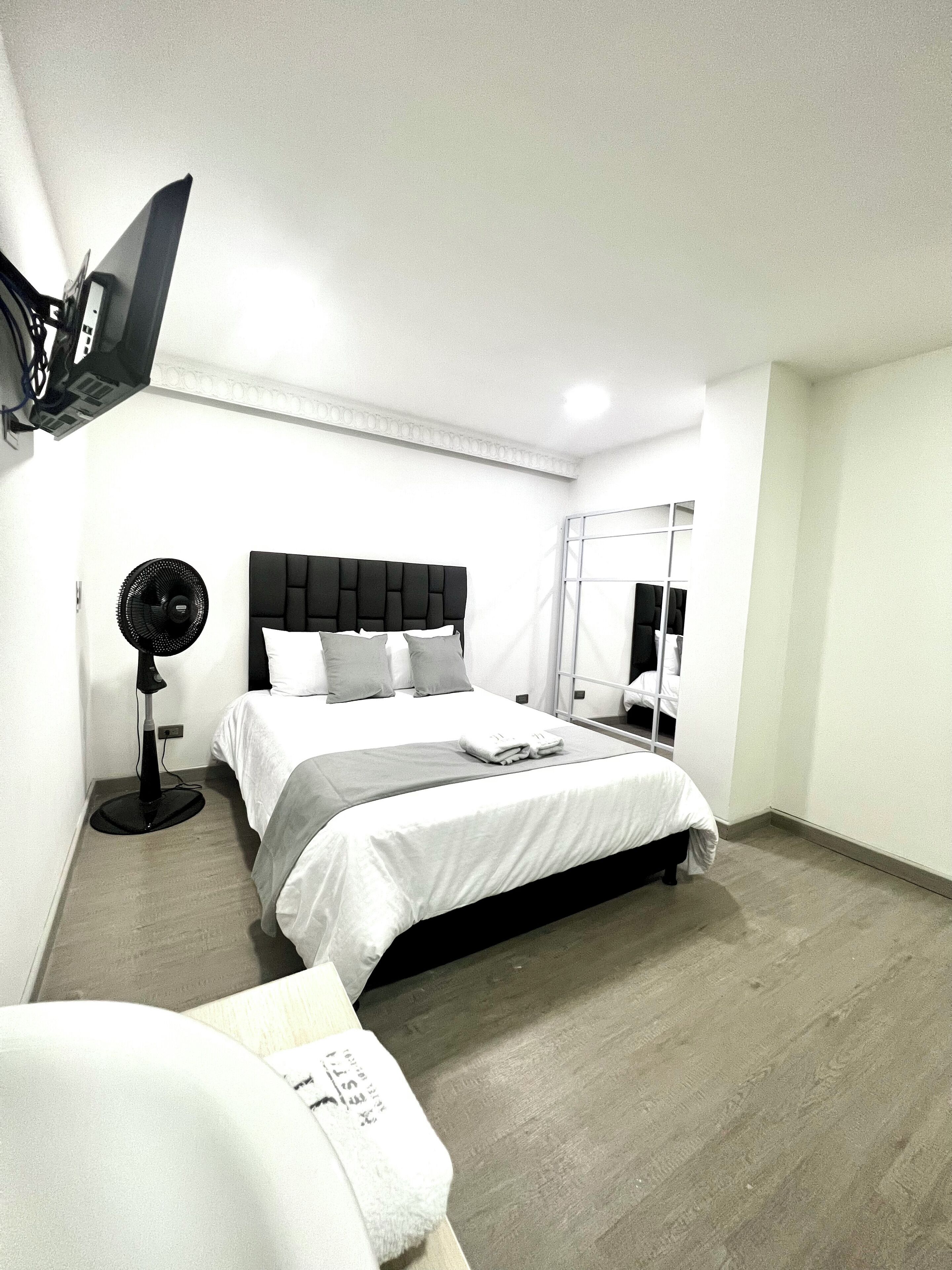 A modern hotel room with a large bed, a standing fan, a flat-screen TV mounted on the wall, and a large mirror.