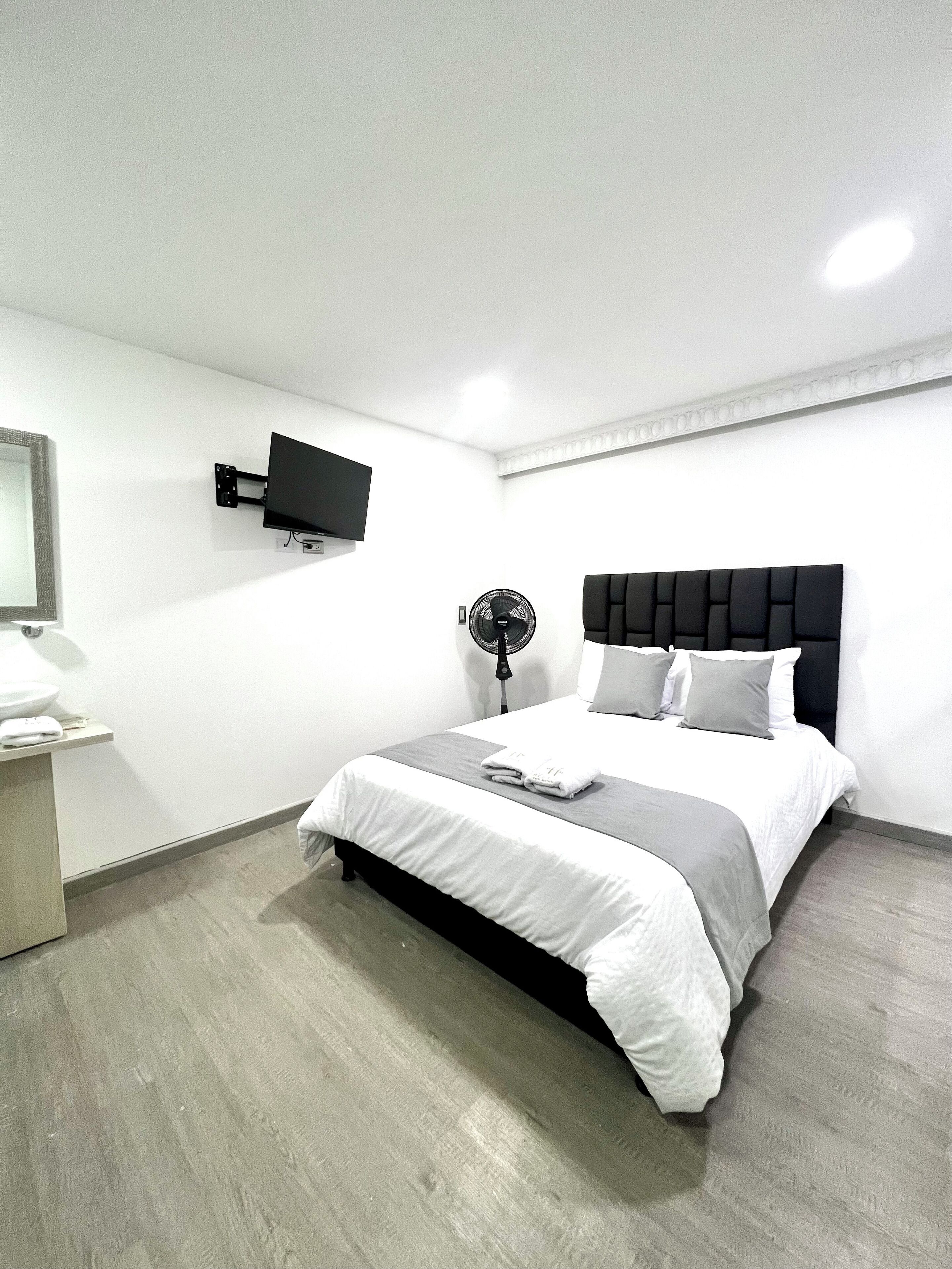 A modern hotel room with a bed, a flat-screen TV mounted on the wall, a bedside table, and a standing fan.