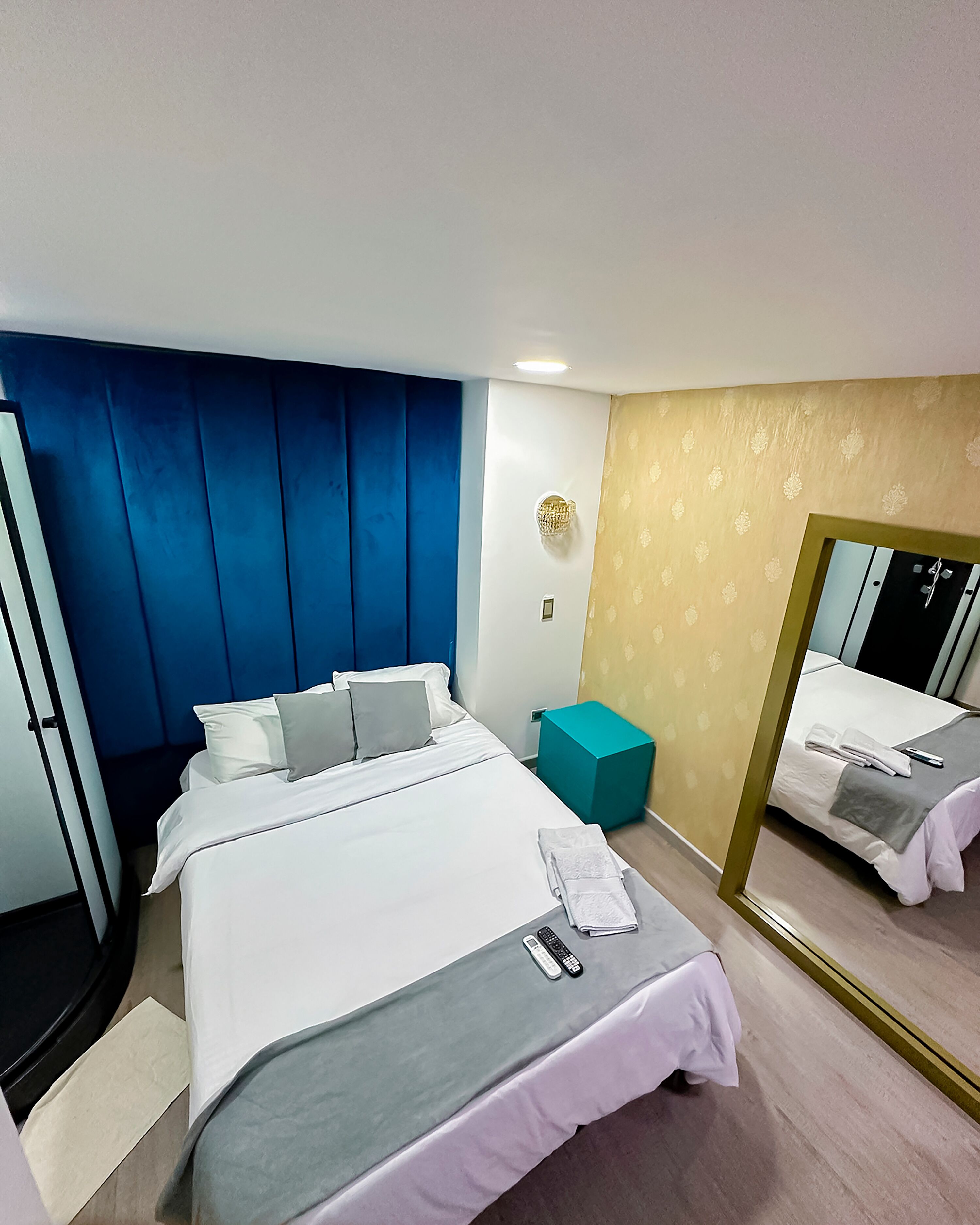 A hotel room with a bed, a small teal nightstand, a mirror, and a wall-mounted light fixture.