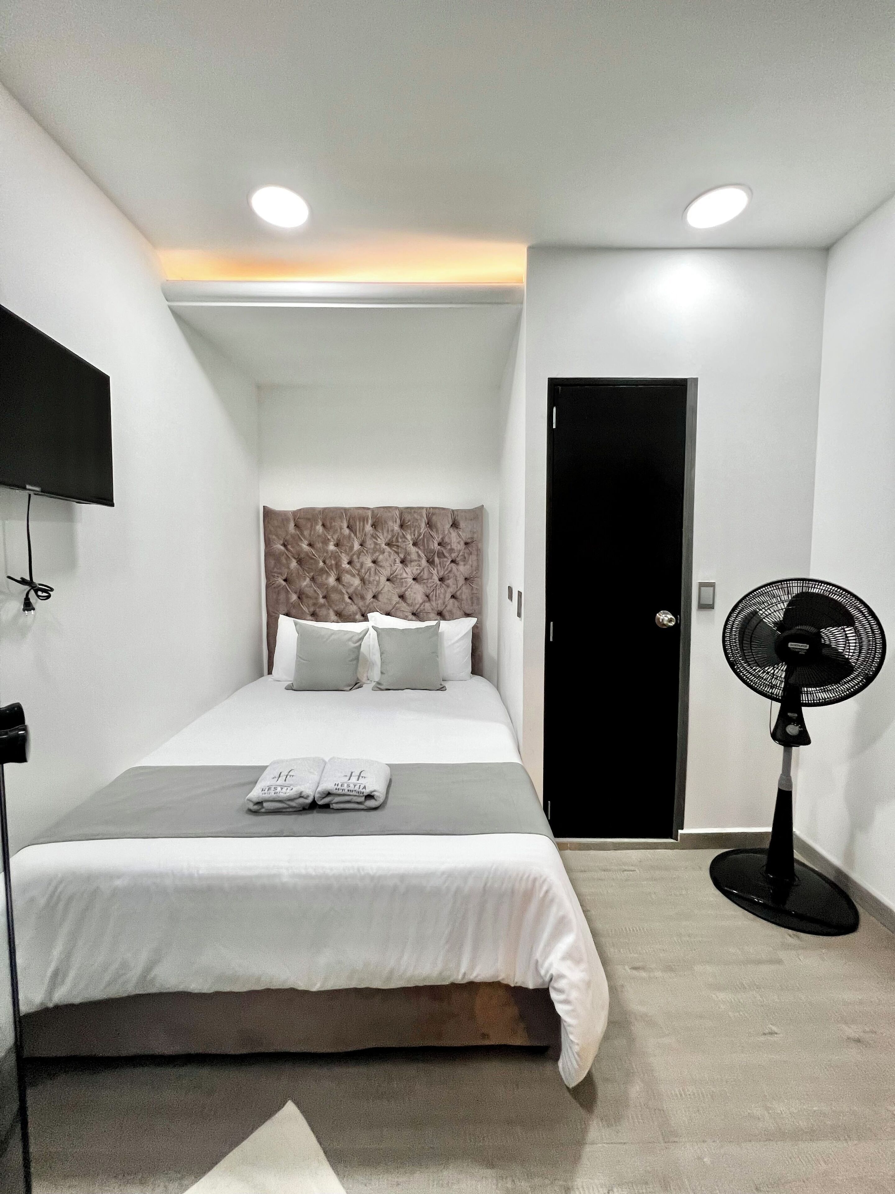 A hotel room with a bed, a flat-screen TV mounted on the wall, a standing fan, and a door.