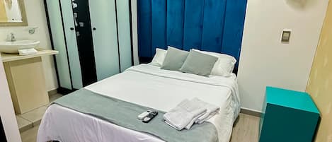 Une chambre d’hôtel moderne comprenant un lit, une salle de bain avec douche, un meuble-lavabo et une table de chevet de couleur turquoise.