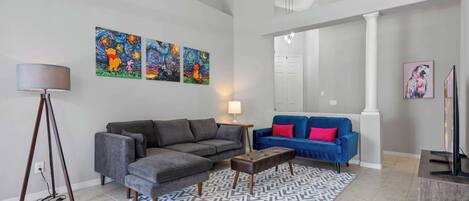 Ein Wohnzimmer mit einem grauen Ecksofa, einem blauen Sofa mit roten Kissen, einem gemusterten Teppich und drei Bildern an der Wand.