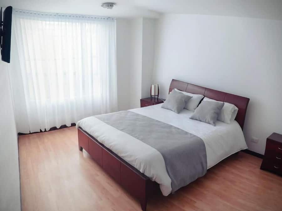 Quarto com cama de madeira, cortinas brancas, mesa de cabeceira e criado-mudo.