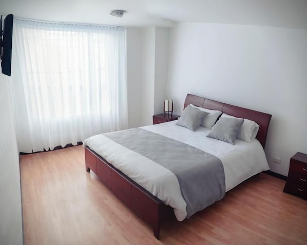 A bedroom with a wooden bed, white curtains, a bedside table, and a nightstand. - Nogal Suite Hotel Pasto (San Juan de Pasto)