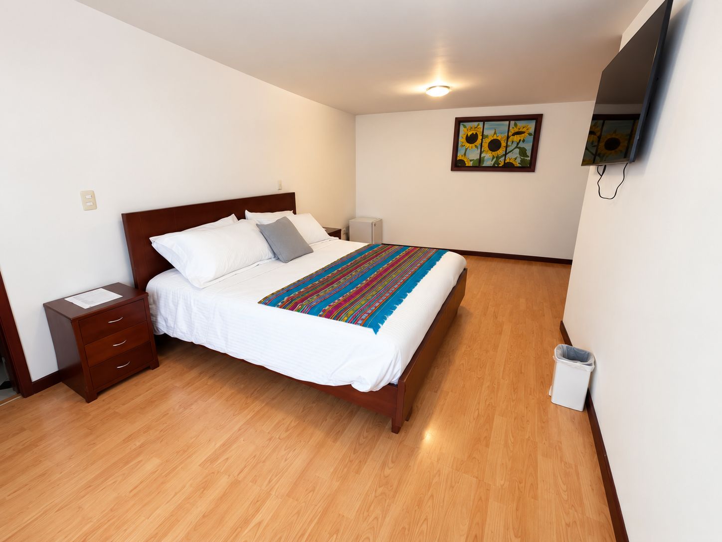 A neatly made bed with a colorful blanket, a wooden nightstand, a small bedside table, a wall-mounted TV, and a painting of sunflowers.