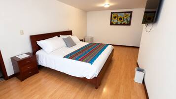 Una cama bien hecha con una manta colorida, una mesita de noche de madera, una pequeña tableta junto a la cama, un televisor montado en la pared y un cuadro de girasoles.