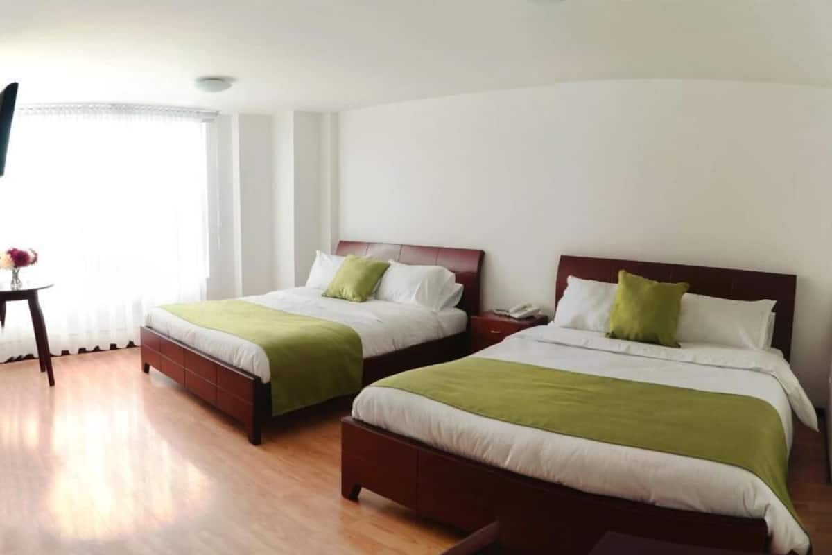 Ett hotellrum med två sängar, var och en med ett träfärgat sänggavel, vita sängkläder och gröna prydnadskuddar. Det finns ett litet bord med en vas med blommor och en platt-TV som är monterad på väggen.