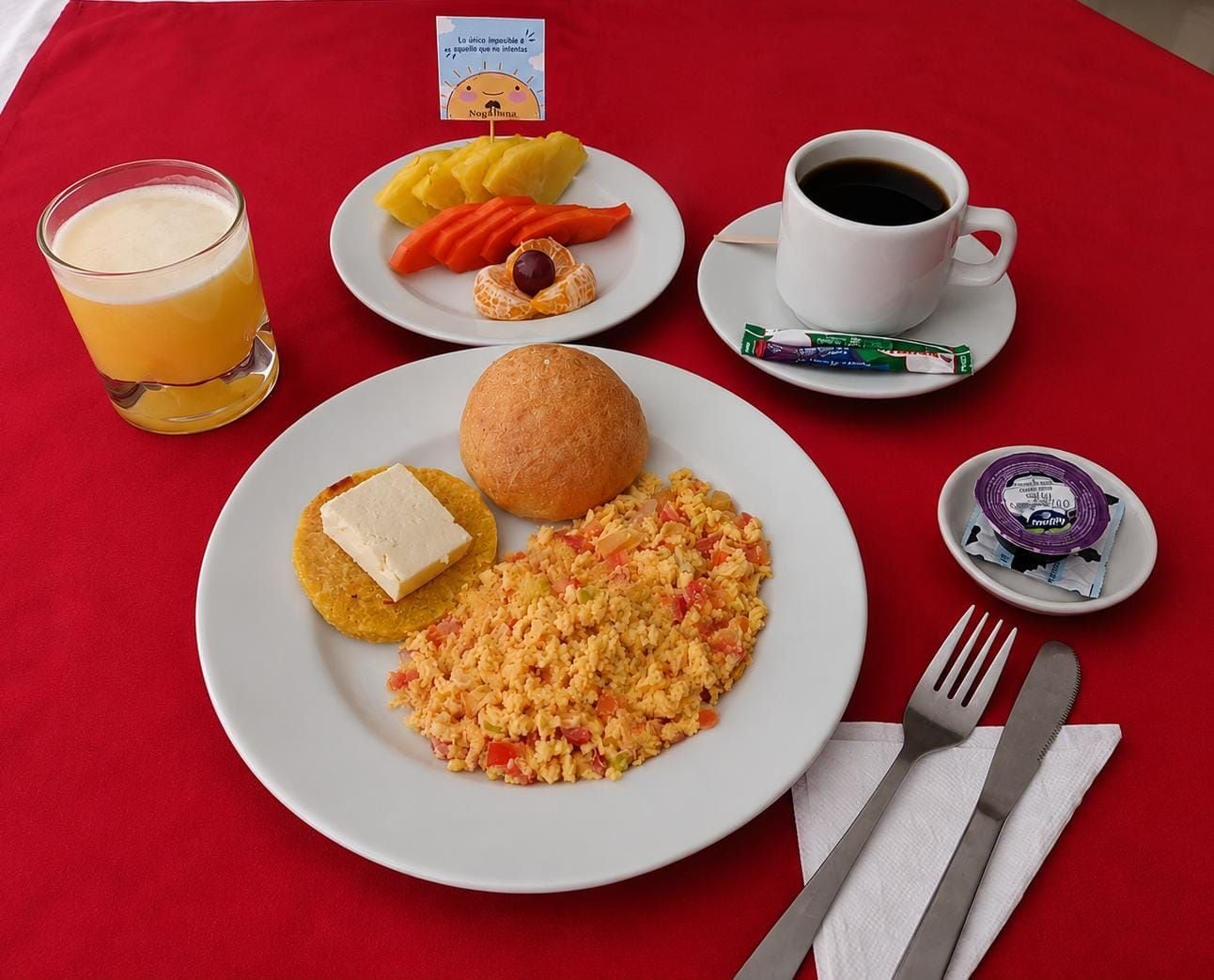 Un desayuno con huevos revueltos, tostadas y un cuenco pequeño de fruta sobre un mantel rojo.