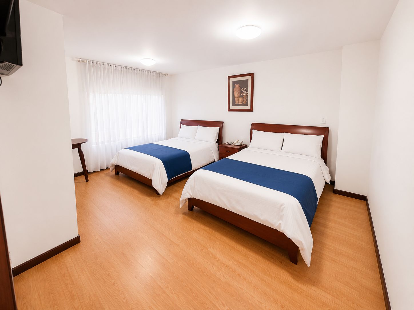 A hotel room with two beds, a bedside table, a nightstand, and a window with curtains.