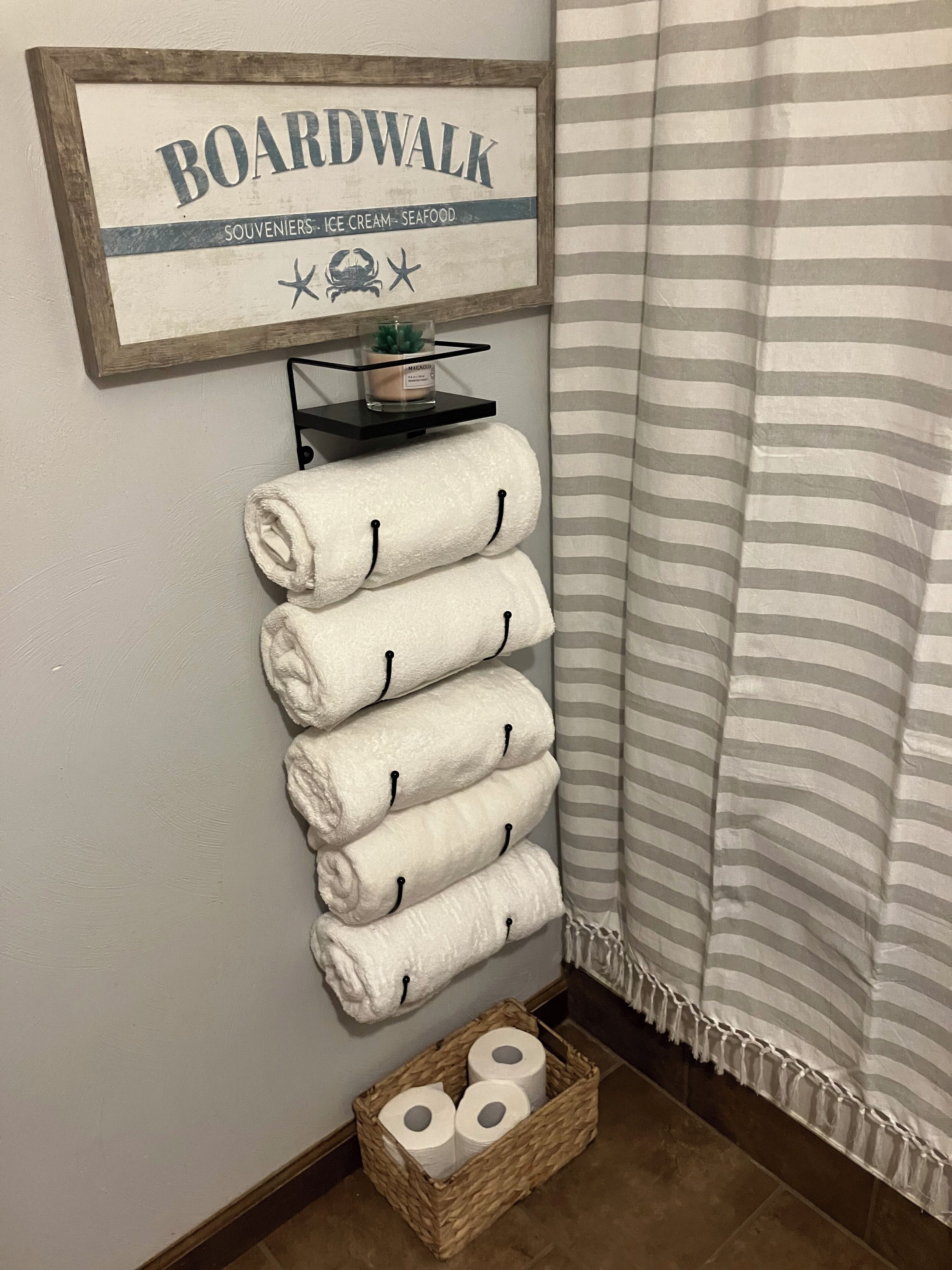 Un baño con un perchero que sostiene cinco toallas blancas, un letrero encima que dice "BOARDWALK SOUVENIERS ICE CREAM SEAFOOD", un estante pequeño con una vela, y una cesta de mimbre con papel higiénico.