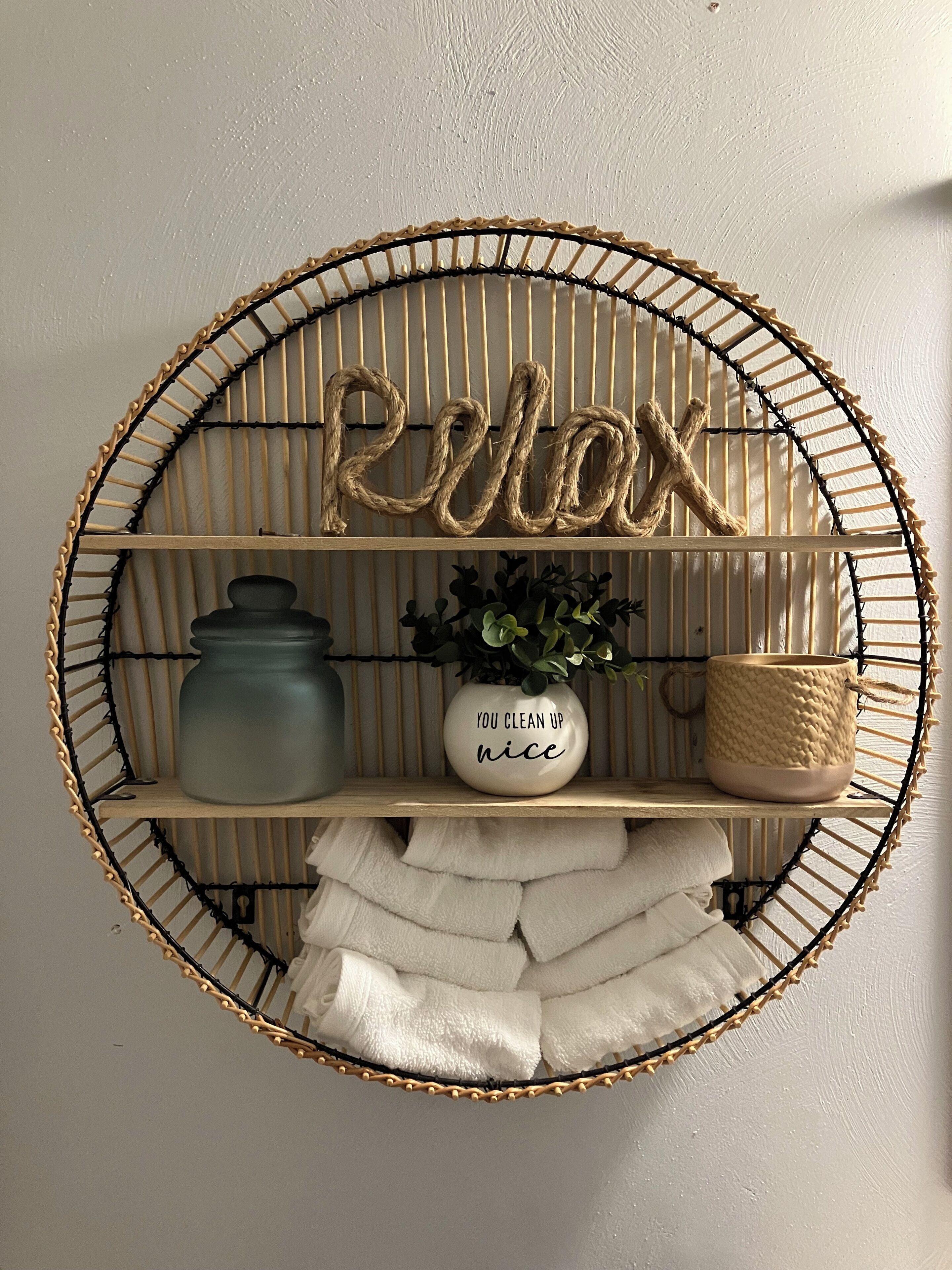 Une étagère murale ornée d’un motif tissé, sur laquelle on trouve une pancarte indiquant « Détendez-vous », une plante en pot, un pot en céramique, un panier tressé et des serviettes soigneusement pliées.
