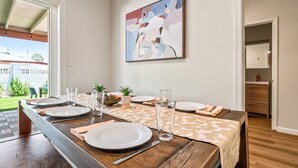 Un comedor con una larga mesa de madera dispuesta para cuatro, un cuadro abstracto grande en la pared y vistas al exterior.