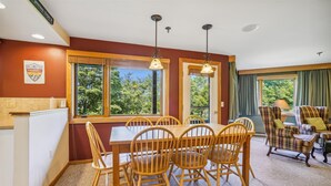 A dining area with a wooden table and chairs, large windows with a view of trees, and a plaid-patterned armchair. - Inviting Two bedroom Two bathroom Highridge Condo B12 (Killington)