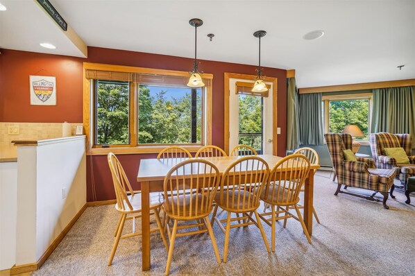 A dining area with a wooden table and chairs, large windows with a view of trees, and a plaid-patterned armchair. - 2-bedroom condo with private sauna, pool, and hot tub. (Killington)