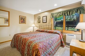 A bedroom with a bed, a window with a view of greenery, a bedside table with a lamp, and a wall with framed pictures. - 2-bedroom condo with private sauna, pool, and hot tub. (Killington)