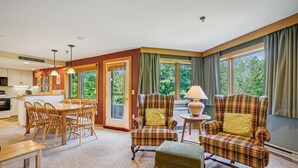 A cozy living room with a dining area, large windows with curtains, and a kitchen in the background. - Inviting Two bedroom Two bathroom Highridge Condo B12 (Killington)