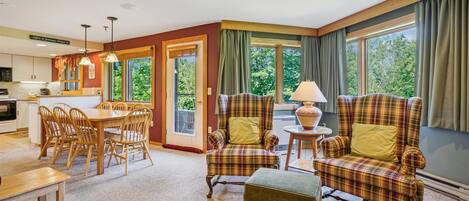 Una sala de estar acogedora con comedor, ventanas amplias con cortinas y una cocina al fondo.