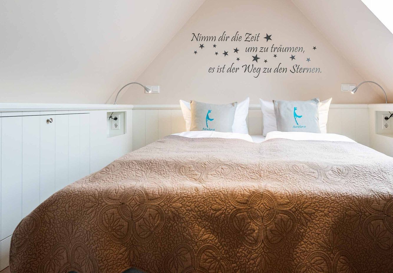 Une chambre avec un grand lit, un mur décoratif représentant un ciel étoilé et un texte en allemand, et une fenêtre laissant entrer la lumière naturelle.