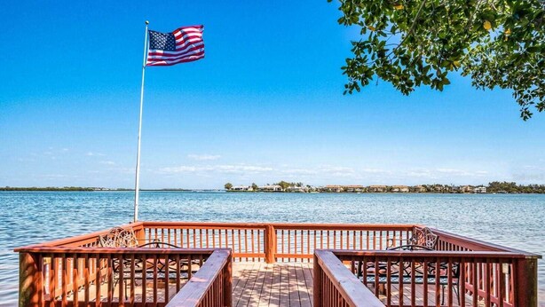 A wooden dock with railings leading to a body of water, an American flag, and a clear view of a distant shoreline.