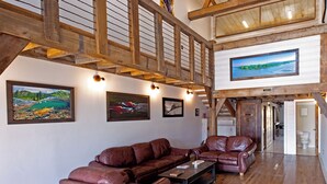 A spacious living area with a large L-shaped leather sofa set, wooden flooring, and a high ceiling with exposed beams.