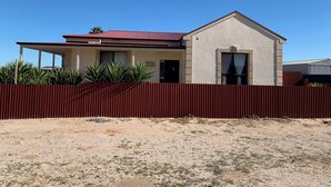 Una casa a due piani con tetto rosso e pareti beige, circondata da una recinzione rossa e un po' di vegetazione.