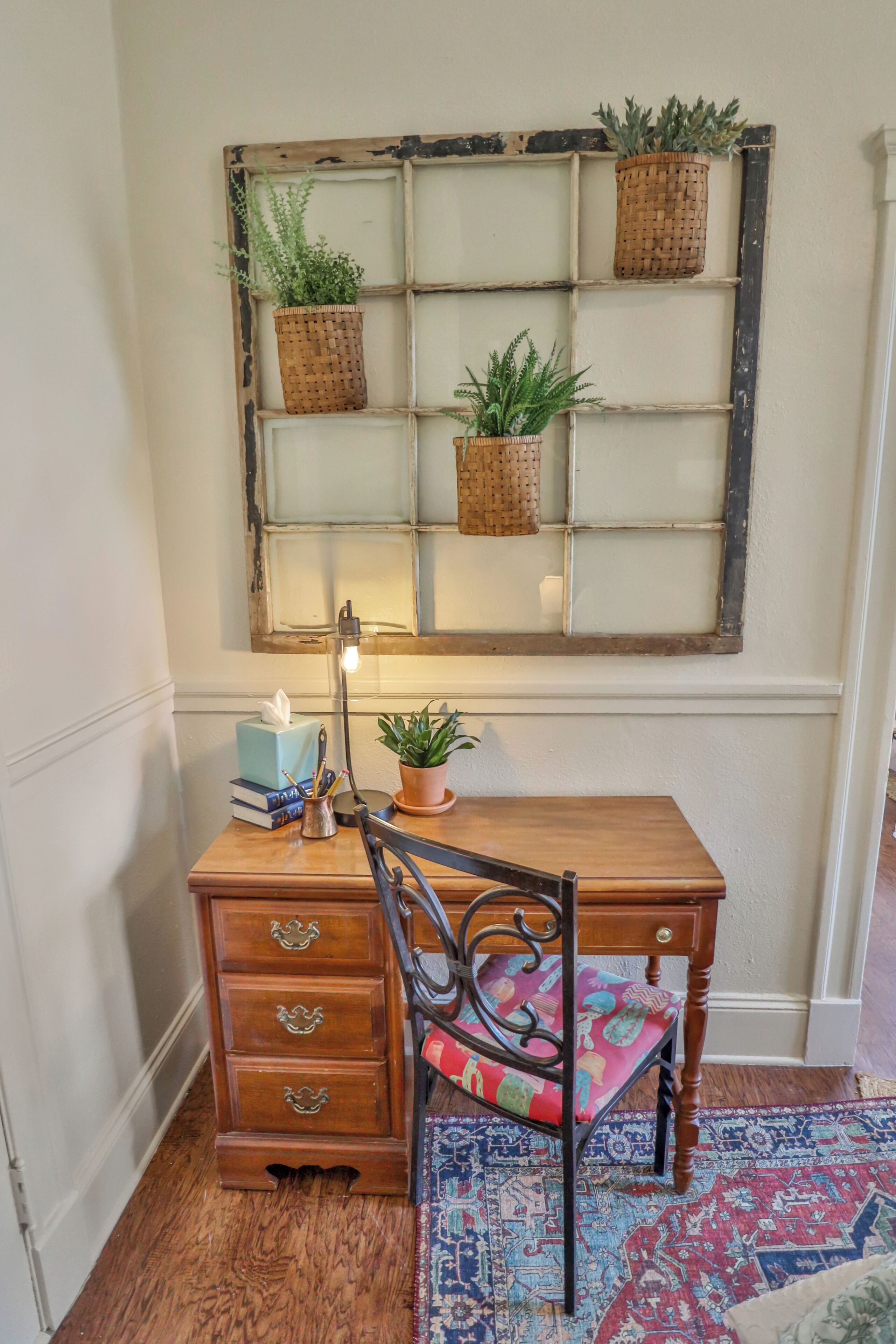 A wooden desk with a chair, a lamp, and a window with plants.