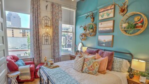 Un dormitorio con una cama, un sillón rojo, una mesita y un ventanal con cortinas.