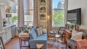 Una sala de estar acogedora con sofá, sillas, una mesita y televisor. Hay cortinas, un ventanal con vistas a edificios y una zona de cocina con microondas y armarios.
