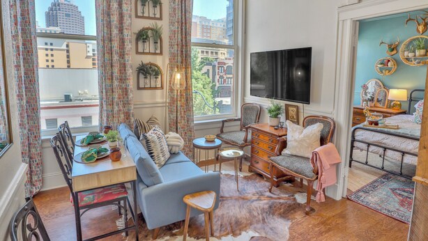 Una sala de estar acogedora con sofá, mesa de comedor y sillas. Hay un televisor montado en la pared y una ventana con cortinas.