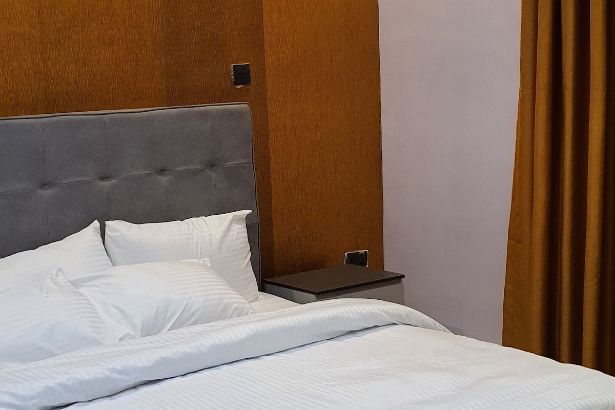 A neatly made bed with white linens in a room with wooden paneling and a brown curtain.