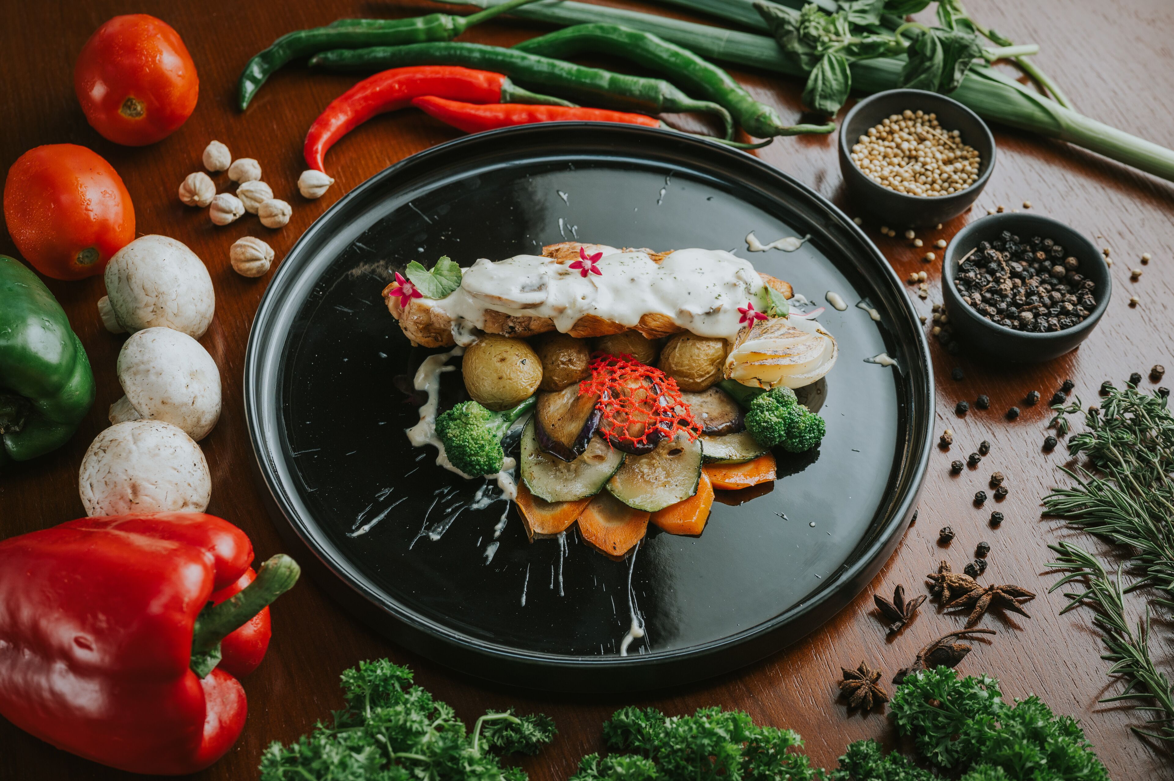 A black plate with roasted vegetables and a creamy sauce, surrounded by fresh herbs and spices.