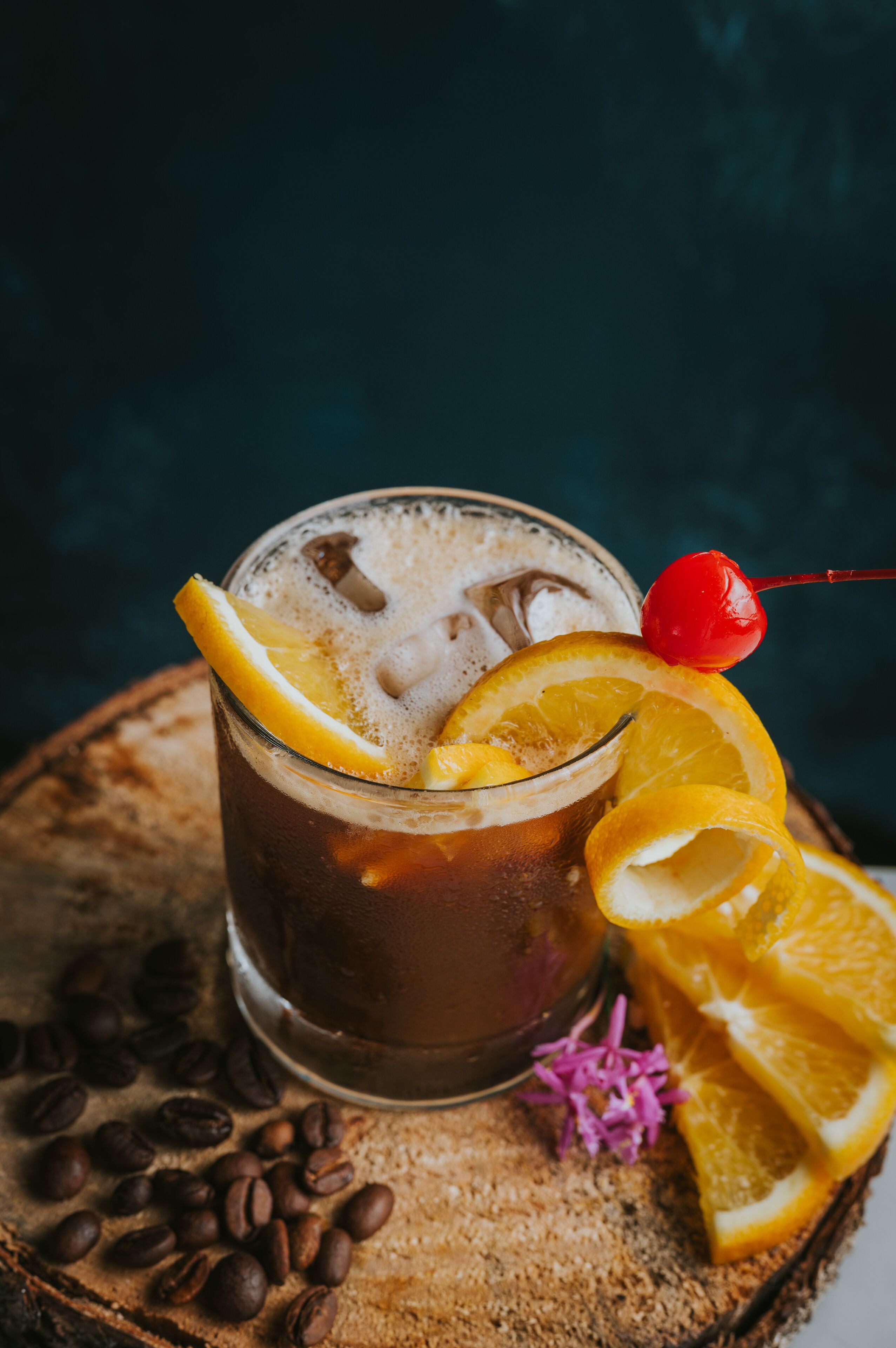 A glass of iced coffee with a cherry on a stick, surrounded by coffee beans and orange slices on a wooden surface.
