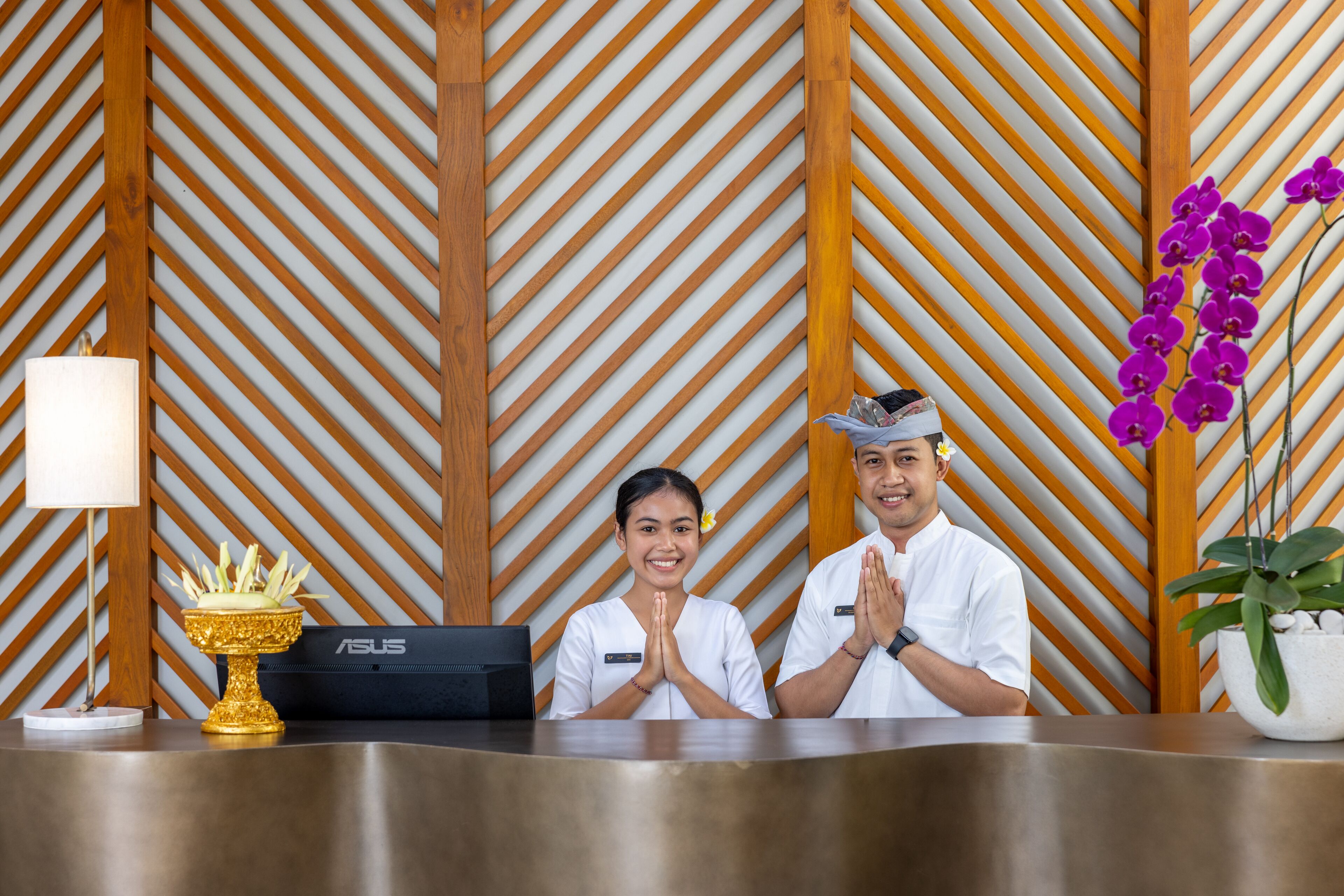 A reception desk with two staff members, a gold decorative item, a plant, and a wall with a diagonal pattern.