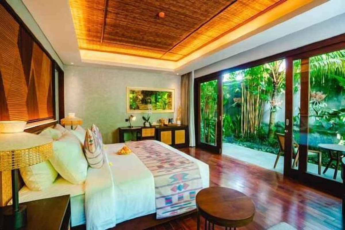 A bedroom with a bed, bedside tables, a desk, and a view of greenery through large windows.