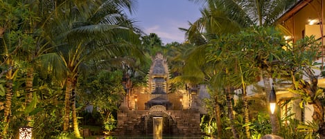 A serene pool area with a stone structure in the center, surrounded by lush palm trees and a well-lit pathway.