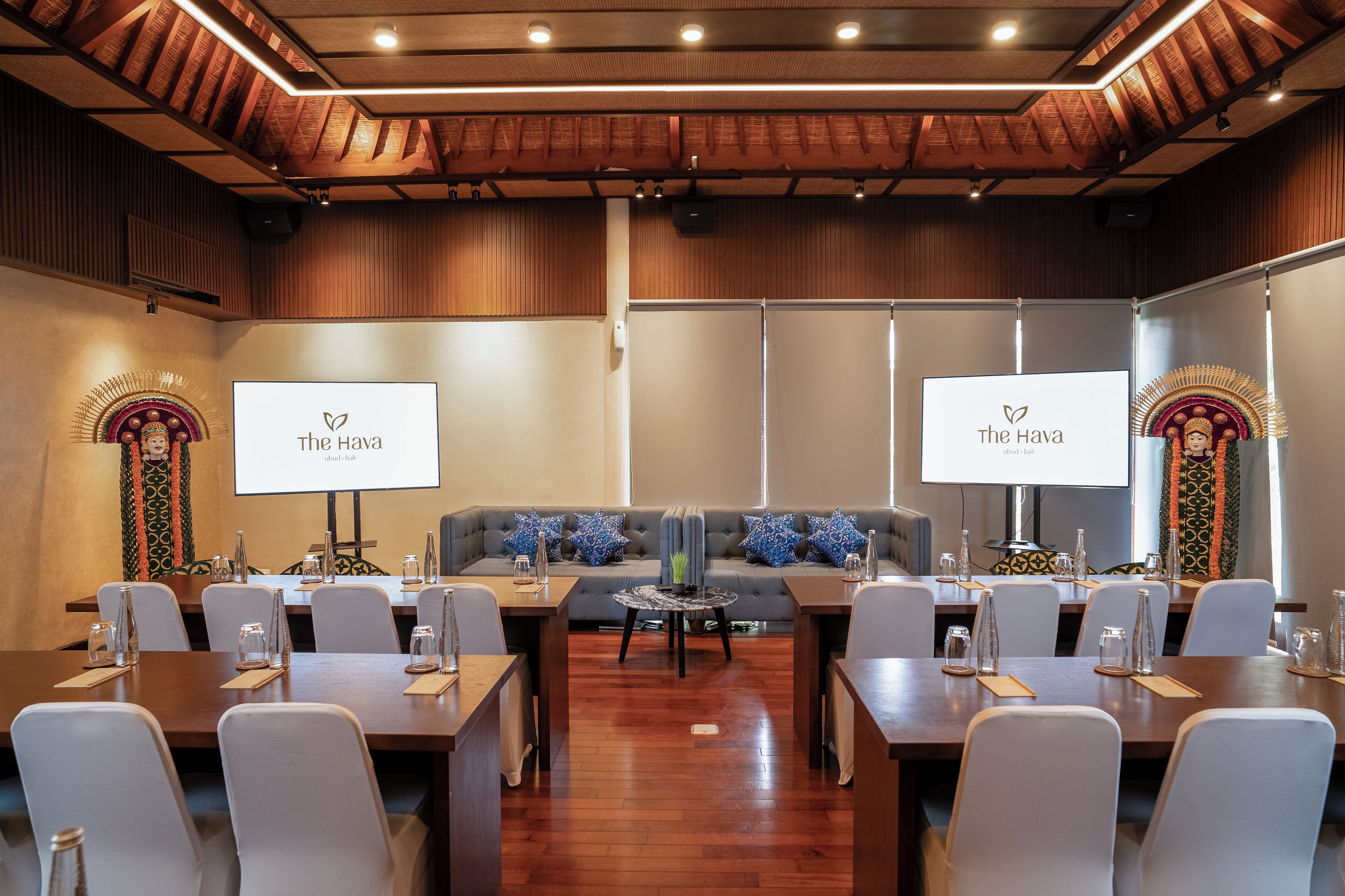 A conference room with a wooden ceiling, two large screens displaying 'The Hava', a grey sofa with blue patterned cushions, and a round table in the center.
