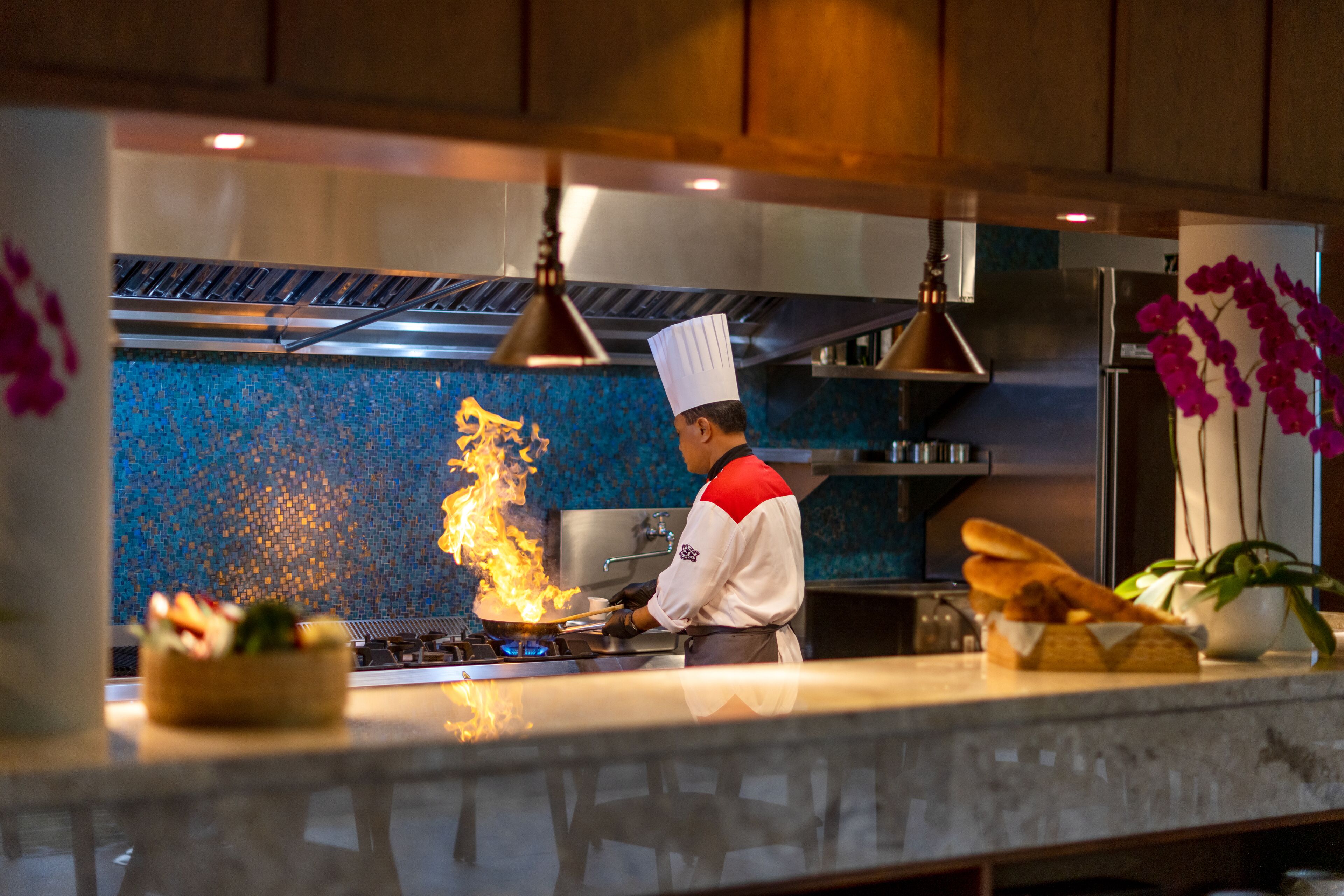 A chef cooking with flames on a stove in a modern kitchen with blue mosaic backsplash and wooden cabinets.