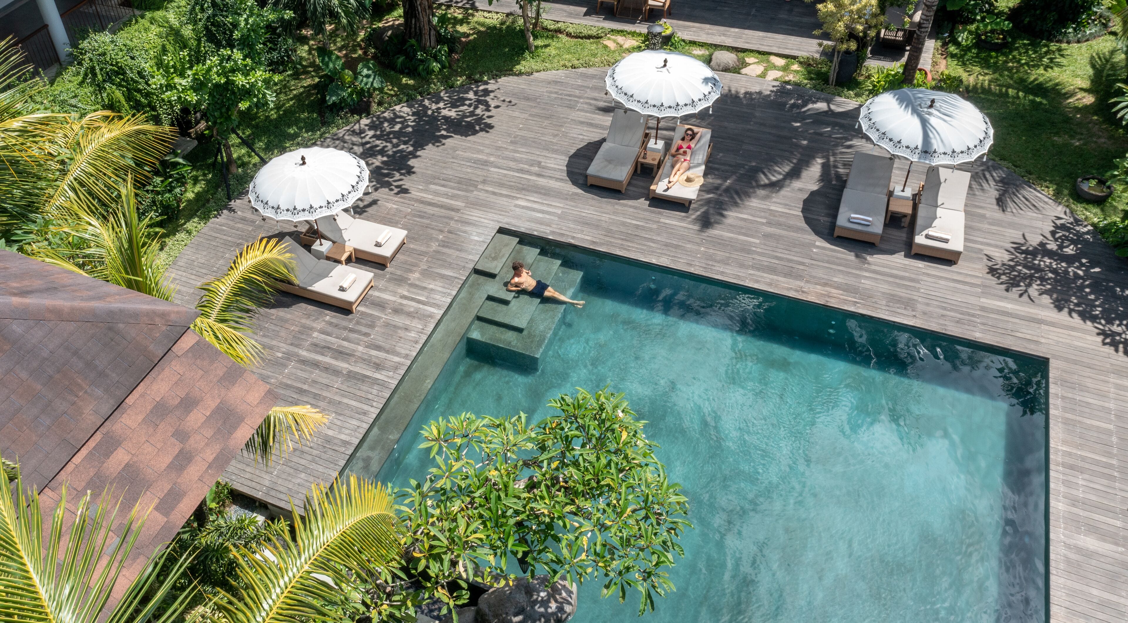 An aerial view of a swimming pool with lounge chairs and umbrellas, surrounded by lush greenery.