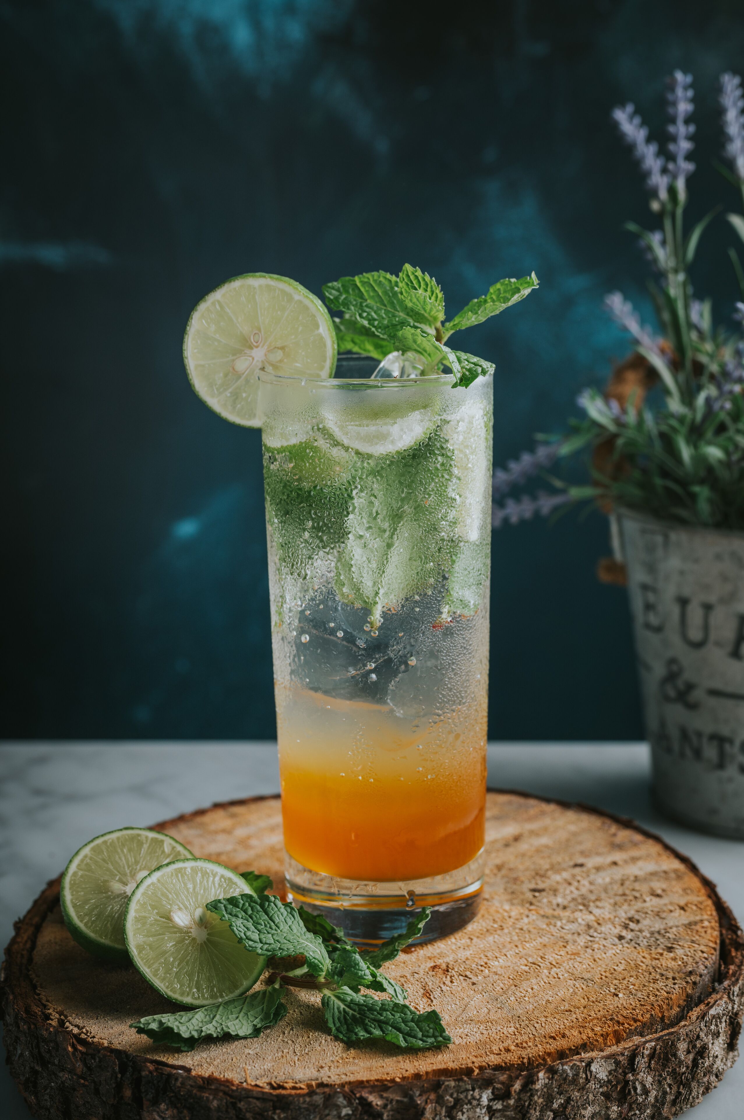 A tall glass of a citrus drink with lime and mint garnish, placed on a wooden slice.