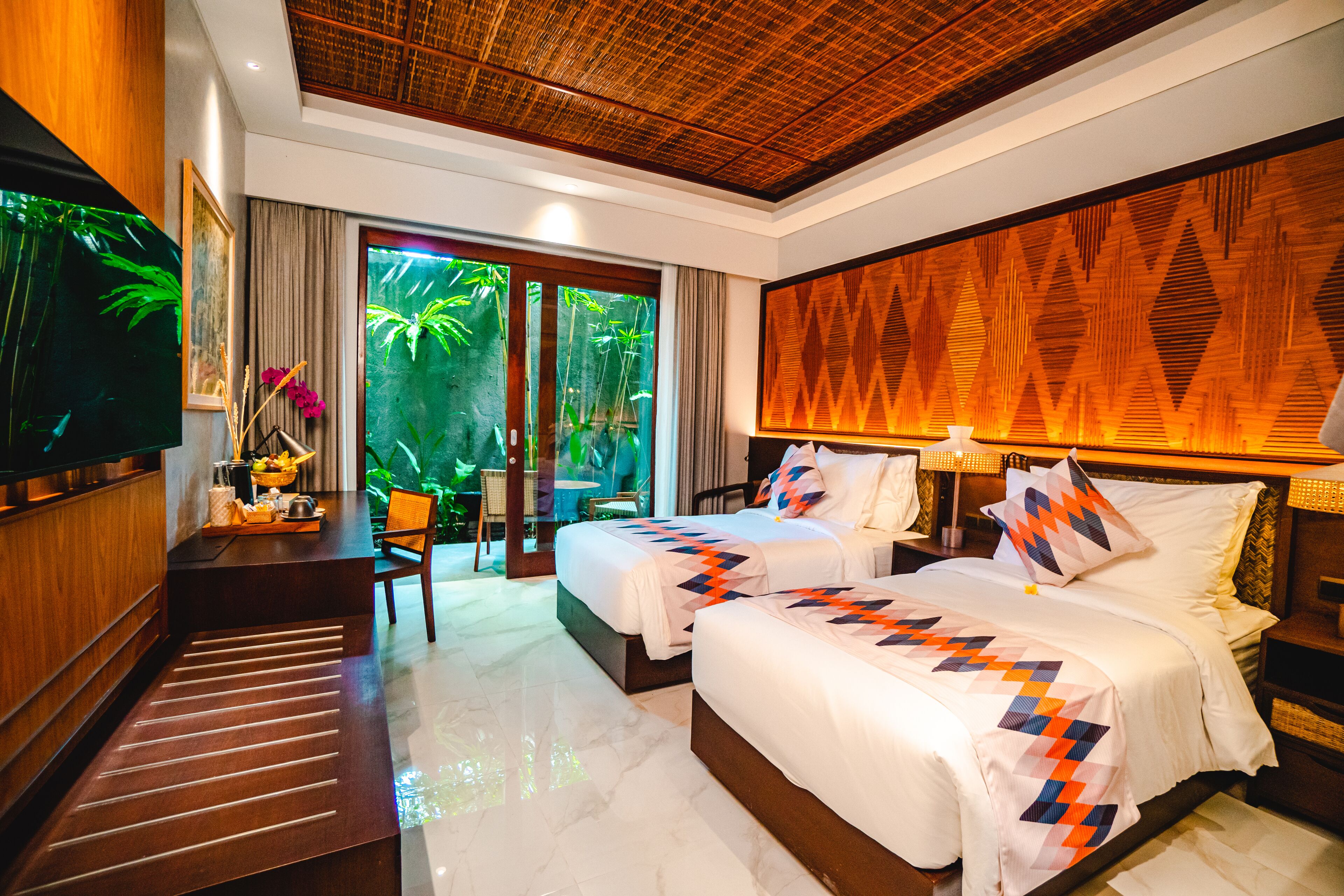 A hotel room with two beds, a TV, a wooden ceiling, and a view of greenery outside.