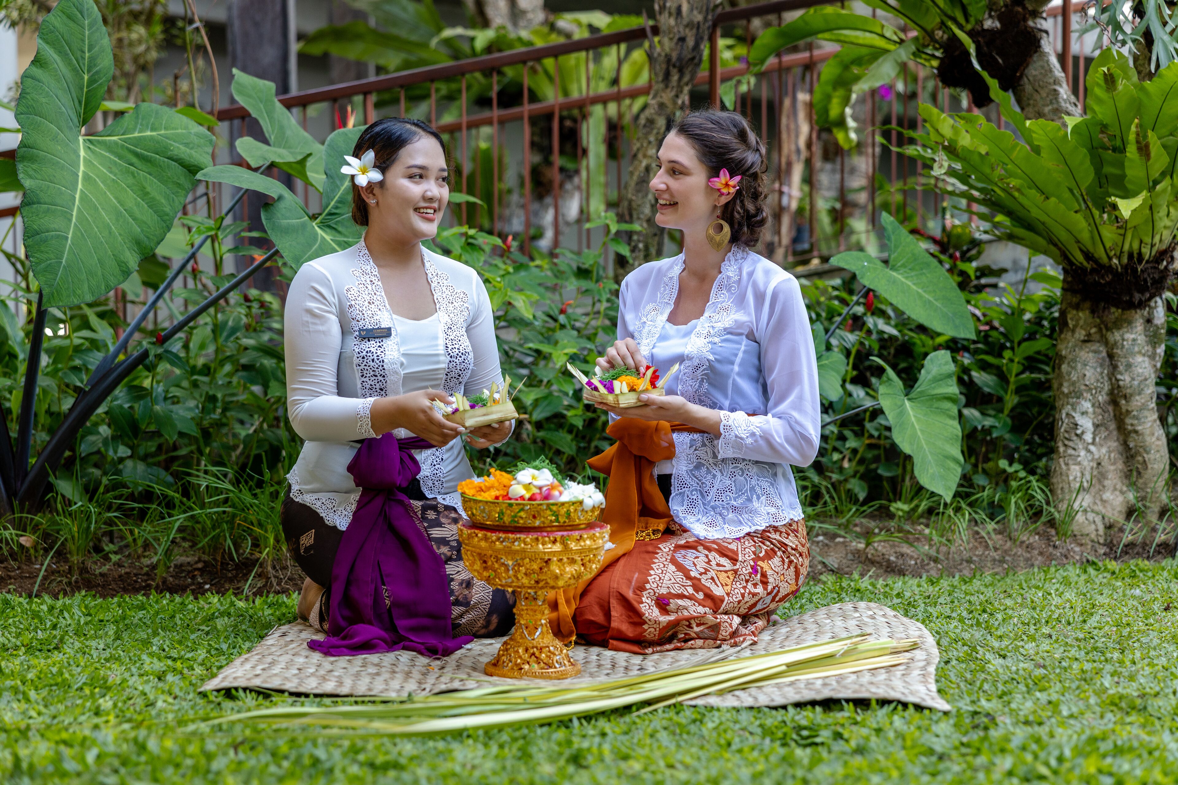 Two individuals seated on a mat outdoors, holding offerings in front of them.