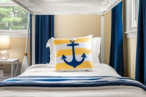 Ein ordentlich bezogenes Bett mit einer blau-weißen gestreiften Tagesdecke, ein Kissen mit Anker-Motiv und eine Lampe auf einem Nachttisch.