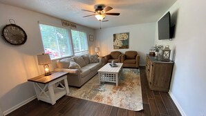 A living room with a sofa, two armchairs, a coffee table, a lamp, a ceiling fan, a clock, and a painting.