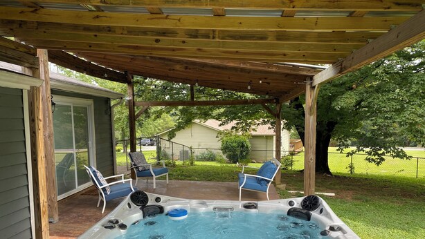 A hot tub under a wooden pergola with two chairs, surrounded by a grassy yard and a house.