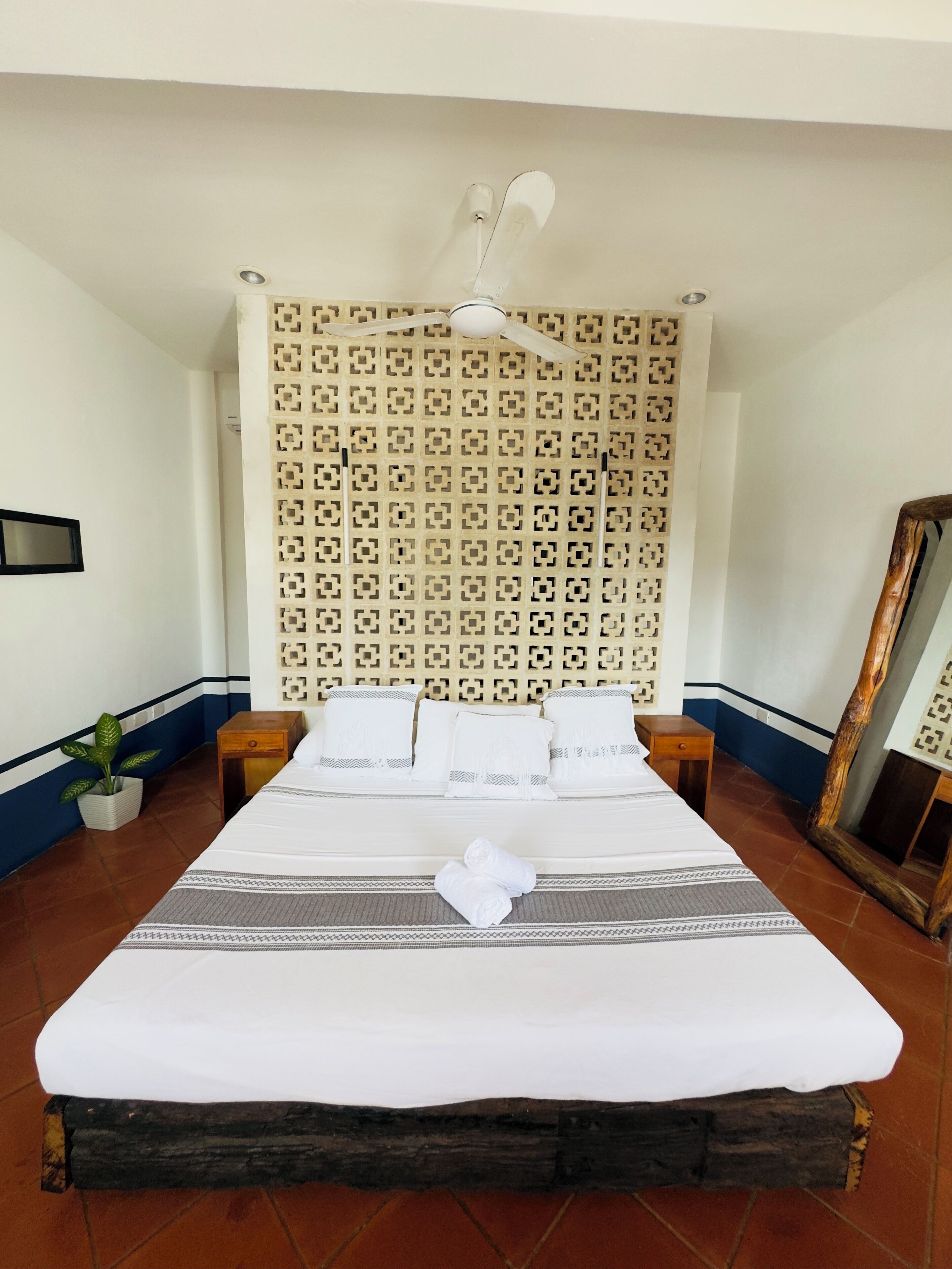 Een net opgemaakt bed met witte lakens en een decoratief kussen, een plafondventilator, een patroonmuur en een spiegel aan de rechterkant.