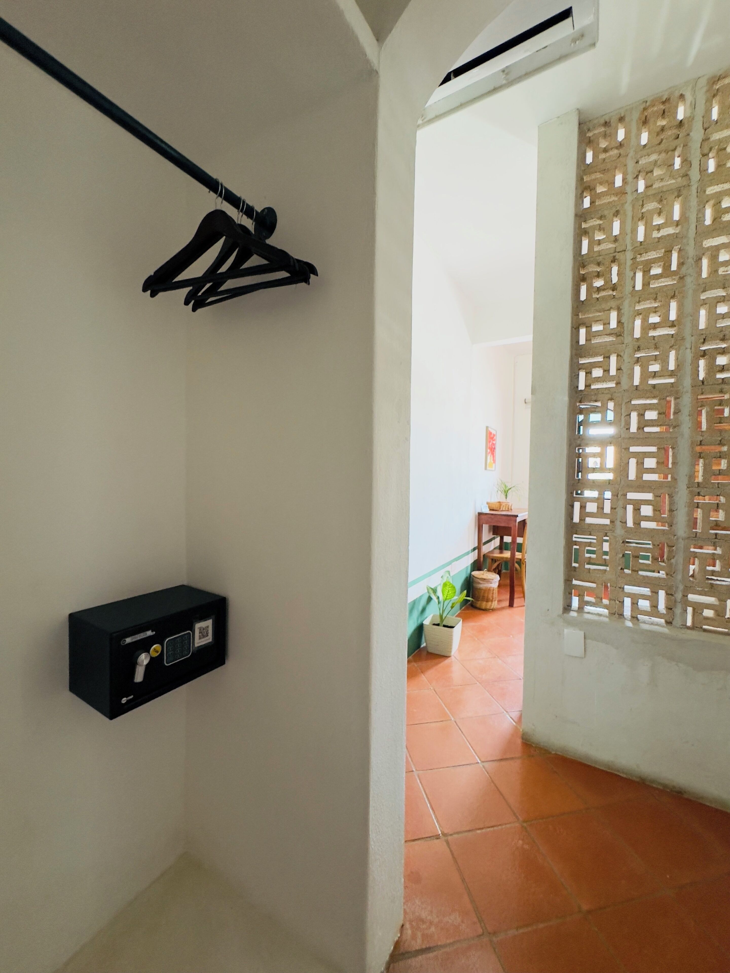 Una habitación con una caja de seguridad negra, un perchero, una ventana con un celosía decorativo y vistas a un comedor con una mesa y sillas.
