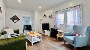 A living room with a green sofa, a blue armchair, a white coffee table, a TV, and a window with floral curtains. - Niseko Riverside by Zen Life : Greenie Cottage (New Glasgow)