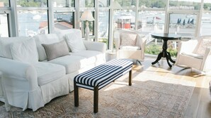 A spacious room with a white sofa, a bench, and two chairs, large windows offering a view of boats and greenery. - Admirals Quarters Inn (Boothbay Harbor)